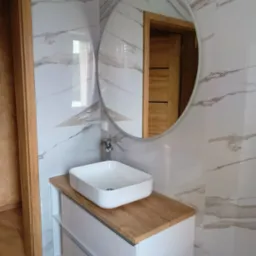 Nowoczesna łazienka po remoncie: biała umywalka nablatowa o kwadratowym kształcie na szafce z drewnianym blatem, okrągłe lustro, płytki imitujące marmur na ścianach, drewnopodobna podłoga.