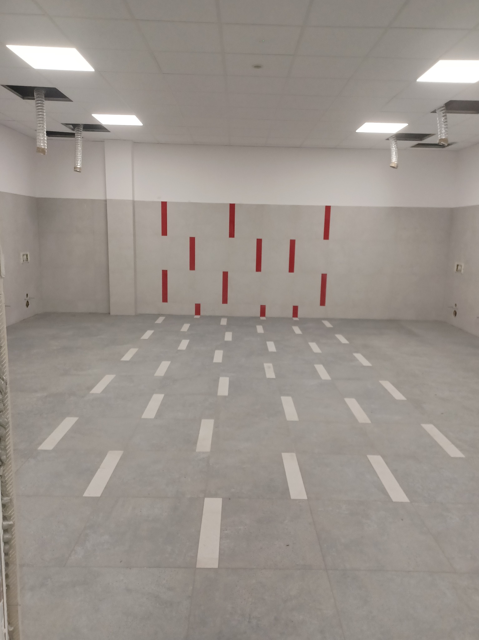 Wykończone pomieszczenie biurowe z szarą podłogą w płytkach z białymi paskami, szare ściany z czerwonymi akcentami, biały sufit z kwadratowymi lampami i widocznymi elementami wentylacji.