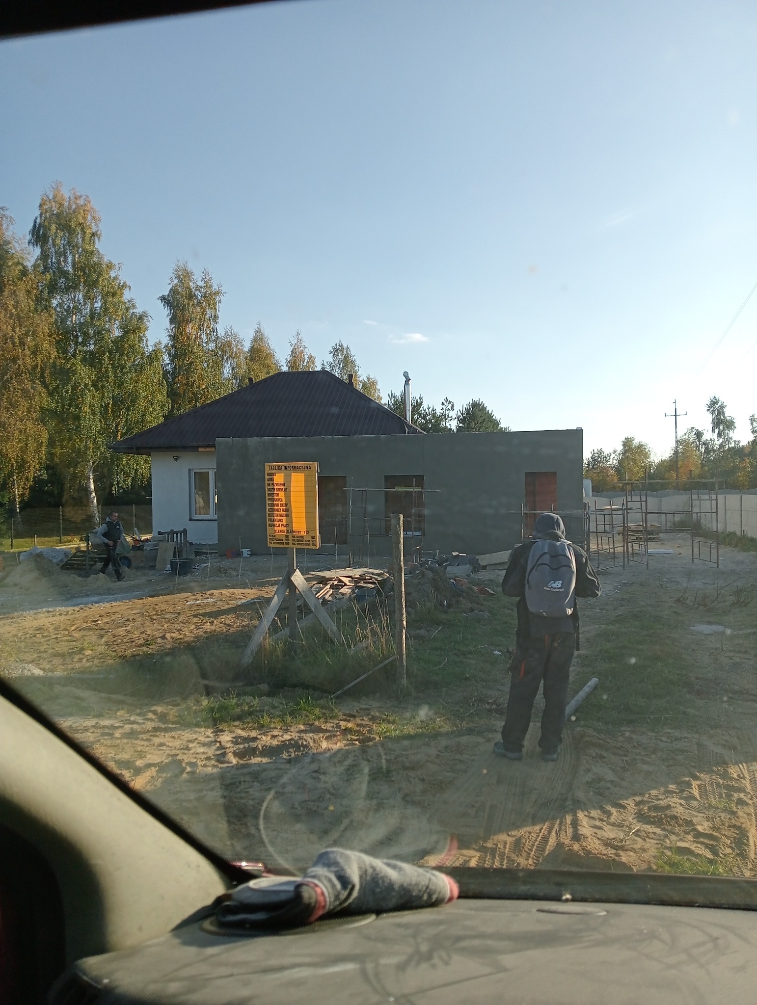 Budynek jednorodzinny w trakcie budowy, widoczna elewacja w szarym tynku, żółta tablica informacyjna budowy oraz pracownik z plecakiem New Balance.