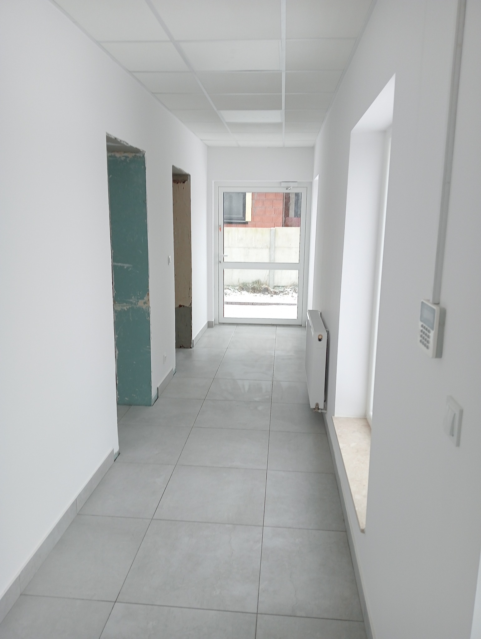 Korytarz w trakcie remontu: położone szare płytki podłogowe, ściany w trakcie malowania, widoczne fragmenty nieotynkowanych futryn drzwiowych, grzejnik i panel alarmowy.