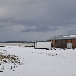 Osoba prywatna - Zimowy krajobraz z budynkami gospodarczymi z czerwonej cegły i kontenerem biurowym, w tle niedokończony budynek mieszkalny i pole pokryte śniegiem, tablice reklamowe z napisem 'Działki...