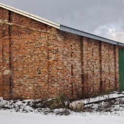 Osoba prywatna - Stary, ceglany magazyn z zielonymi, metalowymi drzwiami w zimowym krajobrazie, częściowo pokryty śniegiem, z widocznymi elementami konstrukcyjnymi i rdzewiejącymi rurami.