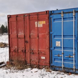 Osoba prywatna - Dwa kontenery magazynowe, czerwony i niebieski, stoją na ośnieżonym terenie. Widoczne ślady użytkowania, rdza na krawędziach. W tle częściowo widoczna konstrukcja budynku w trakcie budowy.