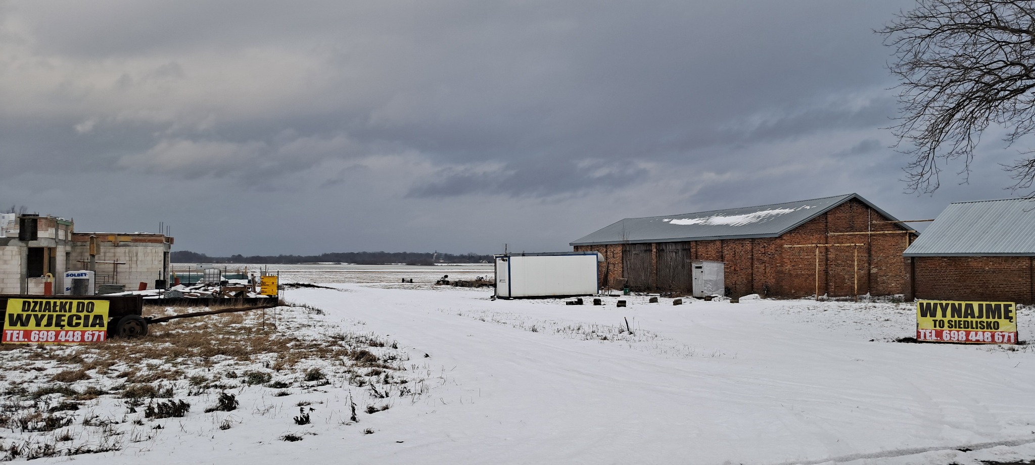 Zimowy krajobraz z budynkami gospodarczymi z czerwonej cegły i kontenerem biurowym, w tle niedokończony budynek mieszkalny i pole pokryte śniegiem, tablice reklamowe z napisem 'Działki...