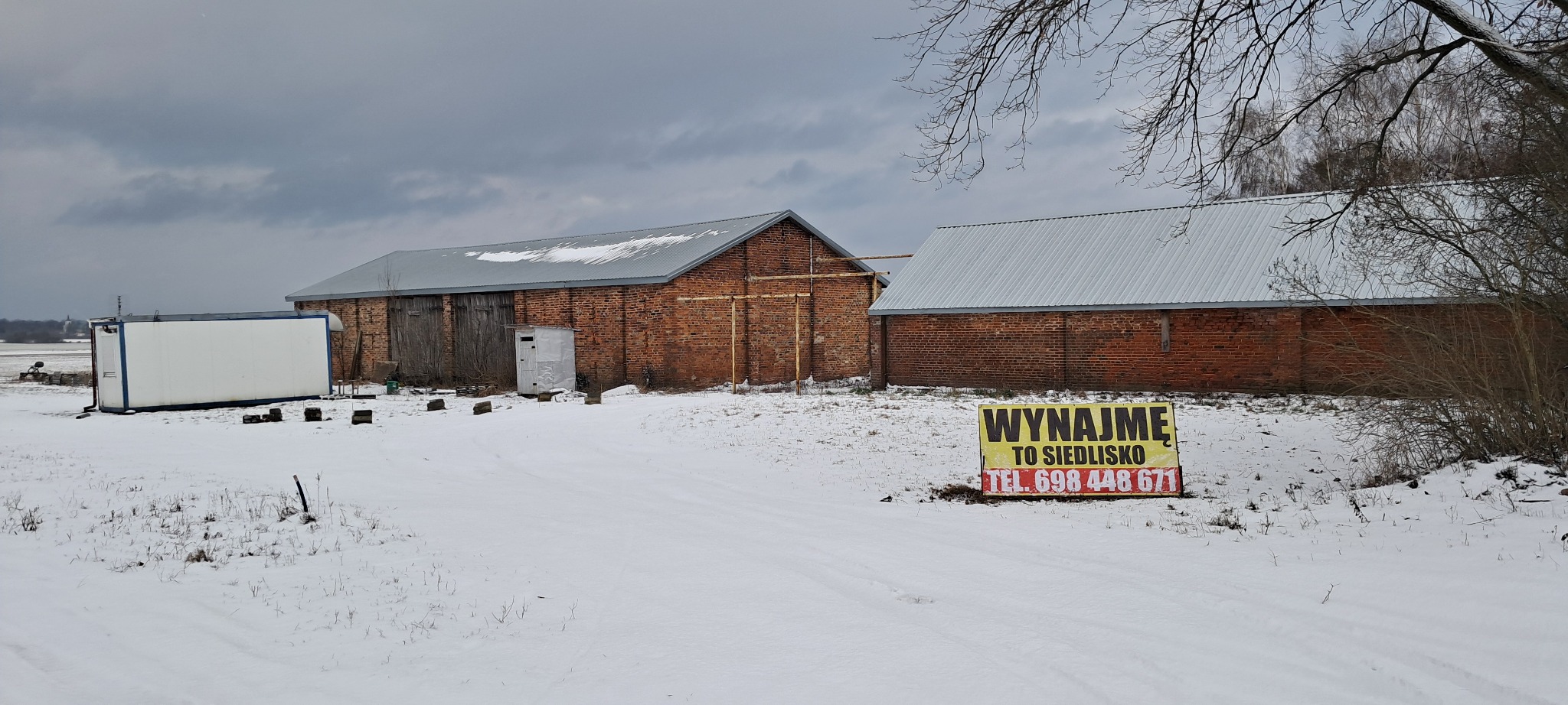 Dwa ceglane budynki gospodarcze z metalowymi dachami, biały kontener i tablica informacyjna z napisem 'Wynajmę to siedlisko' stojące na ośnieżonym terenie pod pochmurnym niebem.