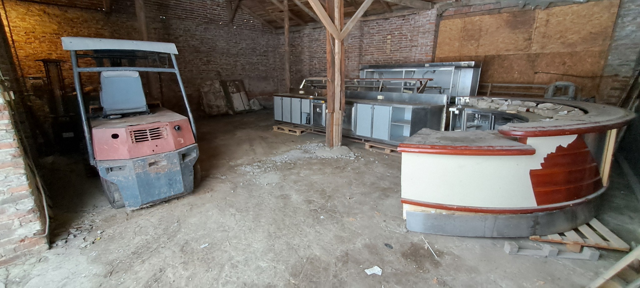 Wnętrze starego magazynu z wózkiem widłowym, metalowymi szafkami i uszkodzonym barem. Widoczne drewniane belki stropowe i ceglane ściany.