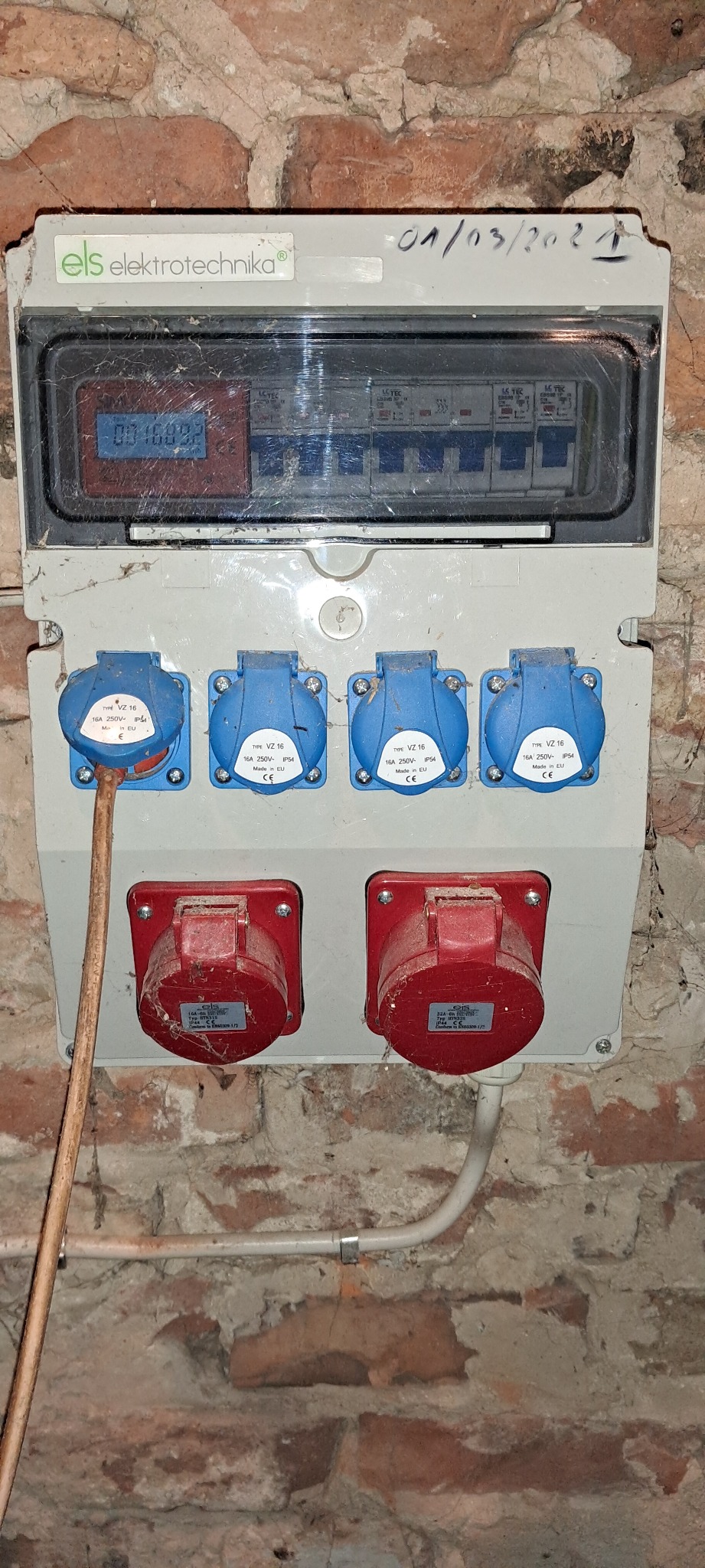 Tablica rozdzielcza z bezpiecznikami i gniazdami siłowymi (16A i 32A) zamontowana na ceglanej ścianie, z widocznym licznikiem energii i datą instalacji (01/03/2021).