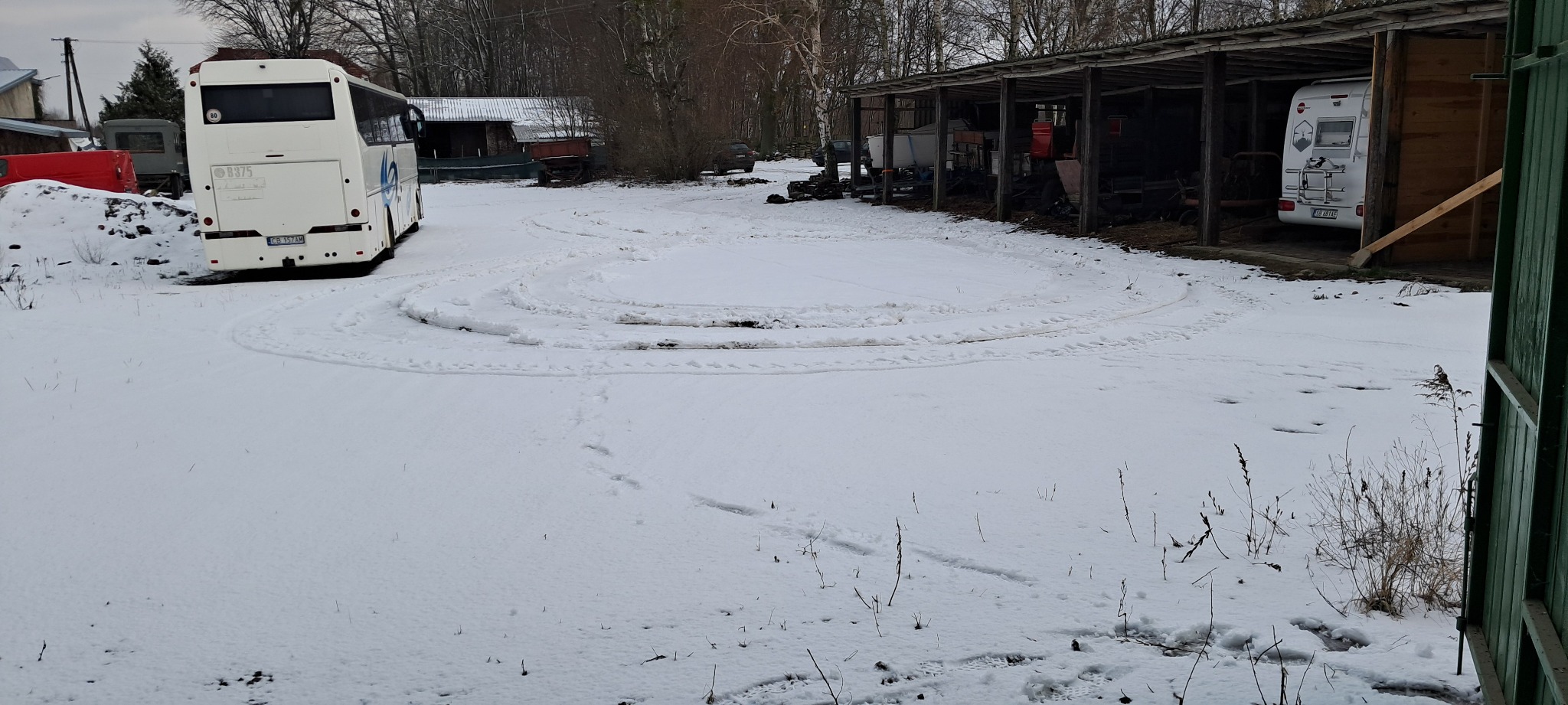 Zimowy plac z zaparkowanym autobusem i kamperem pod zadaszeniem, widoczne ślady opon na śniegu.