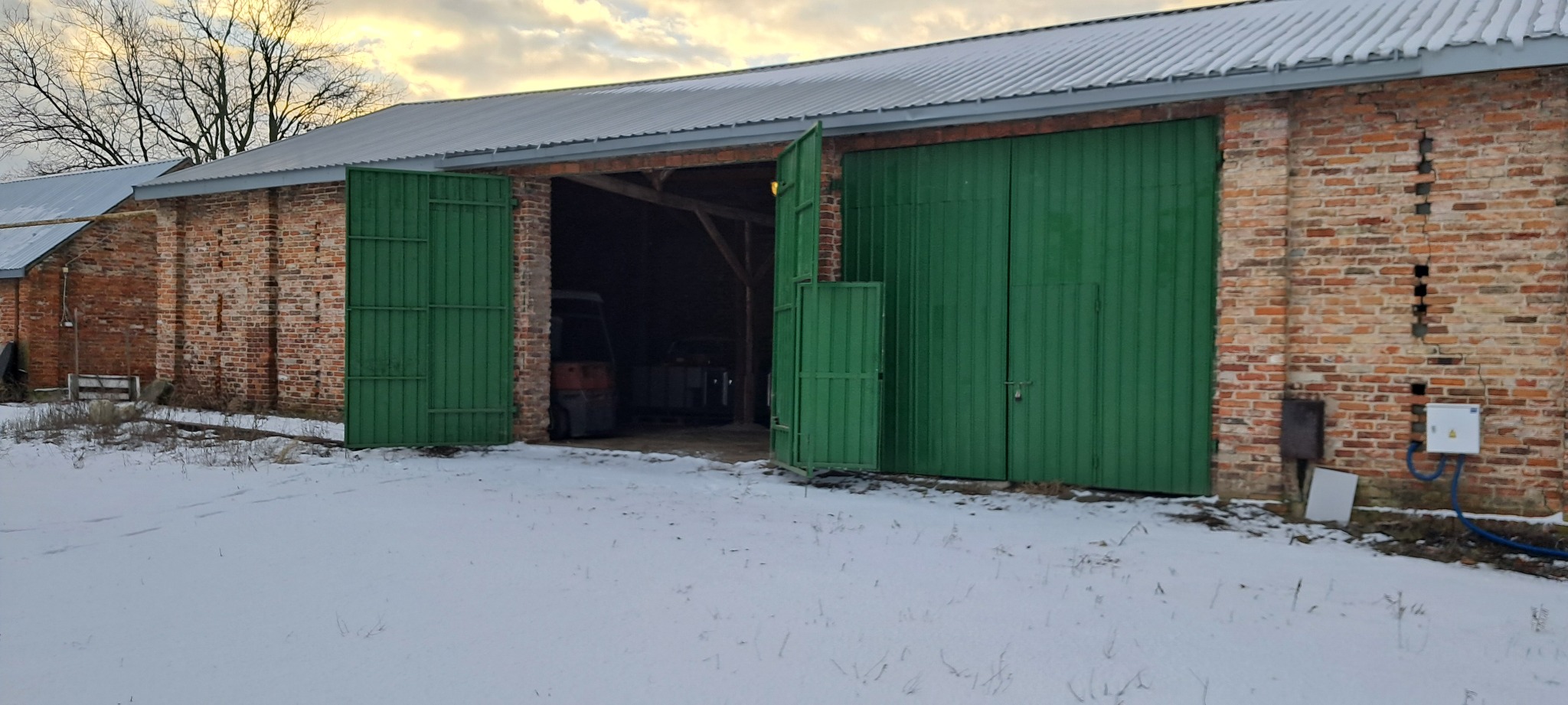 Ceglany budynek magazynowy z otwartymi, zielonymi wrotami, wewnątrz widoczny wózek widłowy, na zewnątrz śnieg.