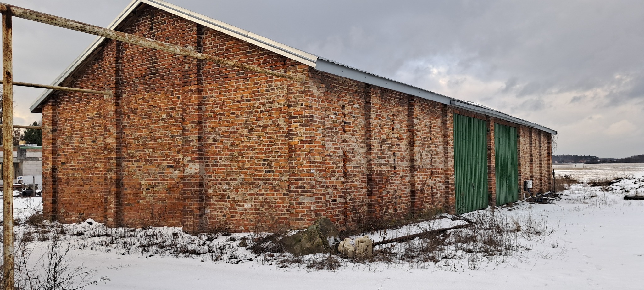 Stary, ceglany magazyn z zielonymi, metalowymi drzwiami w zimowym krajobrazie, częściowo pokryty śniegiem, z widocznymi elementami konstrukcyjnymi i rdzewiejącymi rurami.