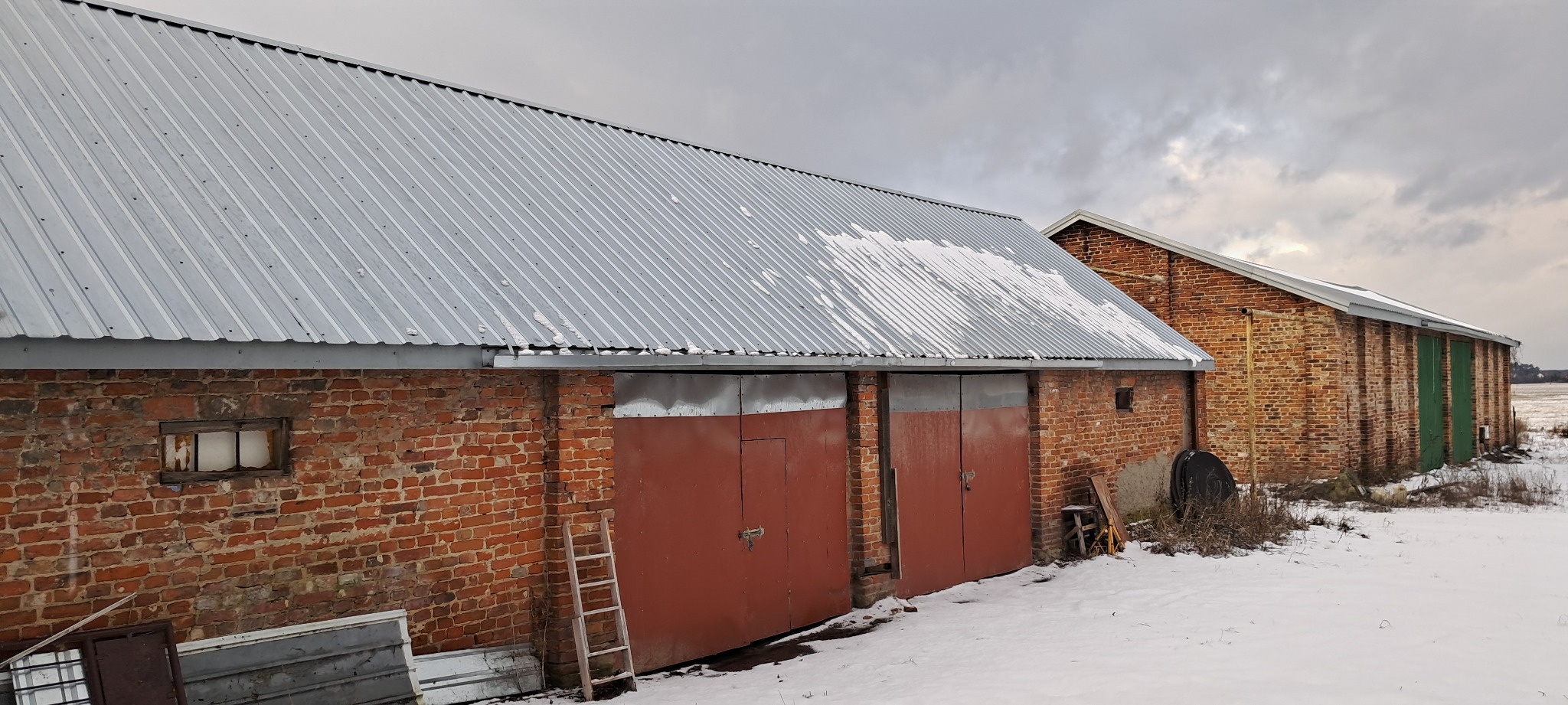 Zewnętrzny widok murowanego magazynu z blachy falistej na dachu, częściowo pokrytego śniegiem, z rdzawymi, zamkniętymi wrotami i drewnianą drabiną opartą o ścianę w zimowej scenerii.