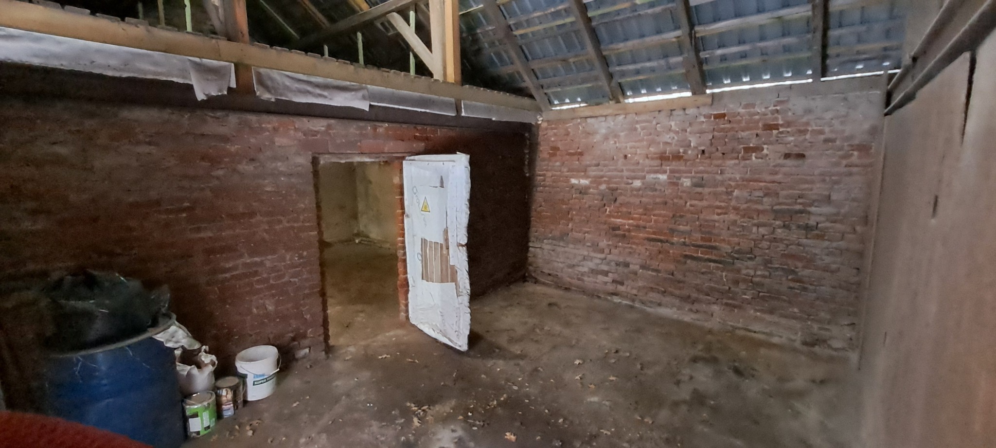 Wnętrze starego, ceglanego magazynu z drewnianą konstrukcją dachu, częściowo odnowione, z niebieską beczką i wiadrami farby w rogu.