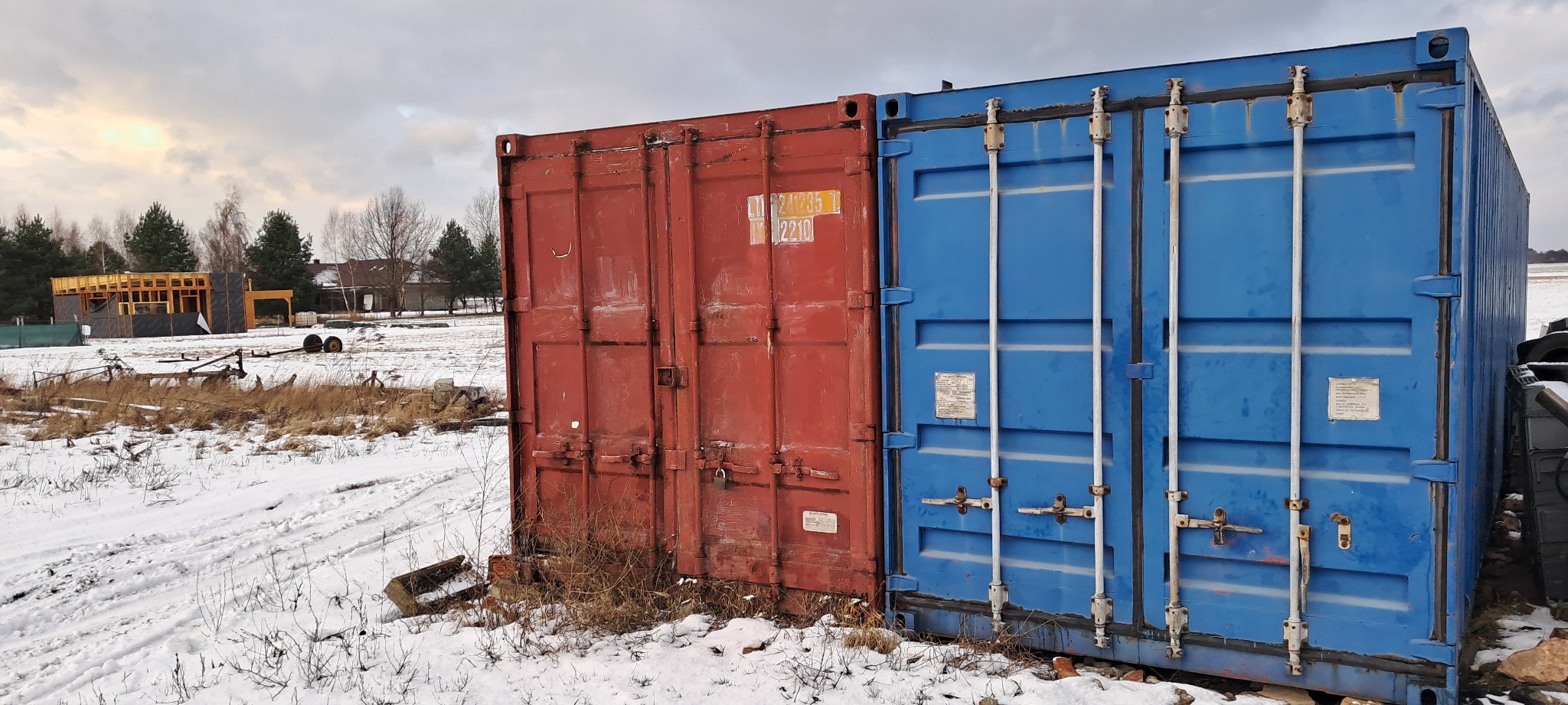Dwa kontenery magazynowe, czerwony i niebieski, stoją na ośnieżonym terenie. Widoczne ślady użytkowania, rdza na krawędziach. W tle częściowo widoczna konstrukcja budynku w trakcie budowy.
