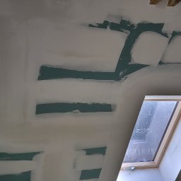 Kacprzak ognobudowlanka - Ściana poddasza z oknem dachowym w trakcie szpachlowania, widoczne pasy zielonej farby i białe wypełnienia szpachlą, fragment rusztowania z czerwoną szmatką w prawym rogu.