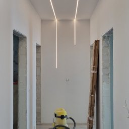 Kacprzak ognobudowlanka - Korytarz w trakcie prac, widoczne oświetlenie ledowe w suficie, przygotowane ościeżnice drzwiowe, odkurzacz przemysłowy i materiały budowlane na podłodze.