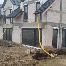 Kacprzak ognobudowlanka - Surowy stan budowy szeregowca z białych bloczków, widoczne żółte węże wentylacyjne wystające z okien, błotniste podłoże, materiały budowlane i biały samochód dostawczy w tle.