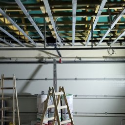 Kacprzak ognobudowlanka - Montaż metalowej konstrukcji podwieszanego sufitu w garażu, widoczne drabiny, materiały izolacyjne i lampa robocza.