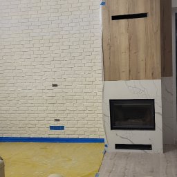 Kacprzak ognobudowlanka - Ściana z cegły pomalowanej na biało, z zainstalowanym nowoczesnym kominkiem obudowanym drewnem i marmurem, podłoga zabezpieczona folią malarską, fragment skórzanej sofy w dolnej części kadru.