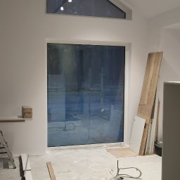 Kacprzak ognobudowlanka - Wnętrze w trakcie wykończenia z dużym oknem zabezpieczonym folią, widoczne listwy przypodłogowe, drabina i narzędzia malarskie na surowej posadzce.