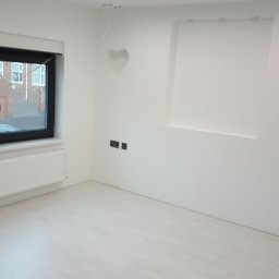 Kacprzak ognobudowlanka - Puste, białe wnętrze po remoncie z jasną podłogą, czarną ramą okienną, białym grzejnikiem, wnęką w ścianie i dekoracyjnym sercem nad gniazdkami.