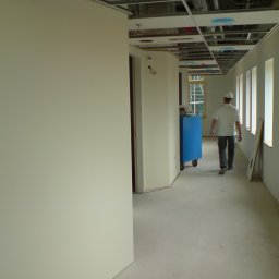Kacprzak ognobudowlanka - Korytarz biurowy w trakcie wykańczania, ściany pomalowane na jasno, widoczne okna, sufit z instalacjami, pracownik w kasku idzie w głąb pomieszczenia.