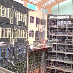 Kacprzak ognobudowlanka - Budynek w trakcie prac elewacyjnych, widoczne rusztowania, okna o nietypowych kształtach i kolorach, ekipa robotników na podnośniku.