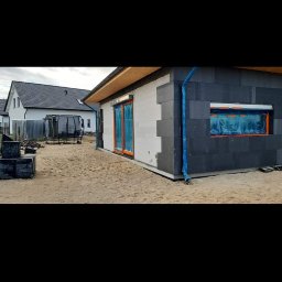 Kacprzak ognobudowlanka - Budowa domu jednorodzinnego z szarych i białych bloczków, z zamontowanymi oknami zabezpieczonymi folią, widoczne są również rury spustowe i trampolina w tle.