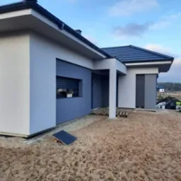 Nowoczesny dom jednorodzinny z elewacją w odcieniach szarości i bieli, widok z zewnątrz, teren wokół domu w trakcie prac wykończeniowych, widoczne palety i materiały budowlane.