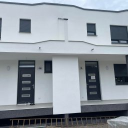 Kacprzak ognobudowlanka - Biała elewacja dwukondygnacyjnego bliźniaka z szarymi roletami i kolumnami, widoczne numery domów 123b i 125, drabina i materiały budowlane leżące przed budynkiem.