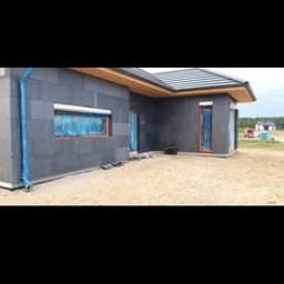 Kacprzak ognobudowlanka - Dom jednorodzinny w trakcie ocieplania elewacji, widoczne szare płyty izolacyjne i zabezpieczone okna folią ochronną, teren wokół domu piaszczysty.