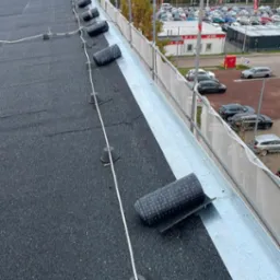 Czarny, płaski dach pokryty papą z widocznymi rolkami papy i elementami mocującymi, w tle budynki i parking.