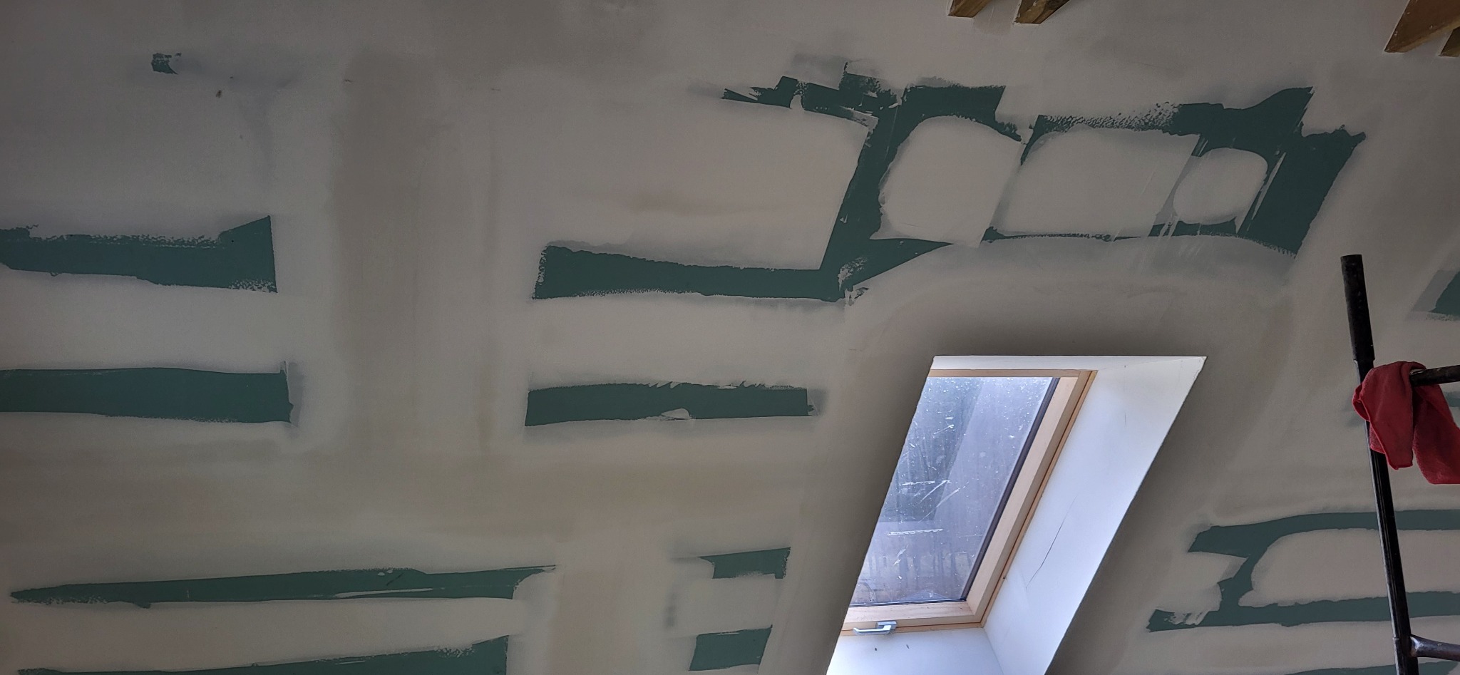 Ściana poddasza z oknem dachowym w trakcie szpachlowania, widoczne pasy zielonej farby i białe wypełnienia szpachlą, fragment rusztowania z czerwoną szmatką w prawym rogu.