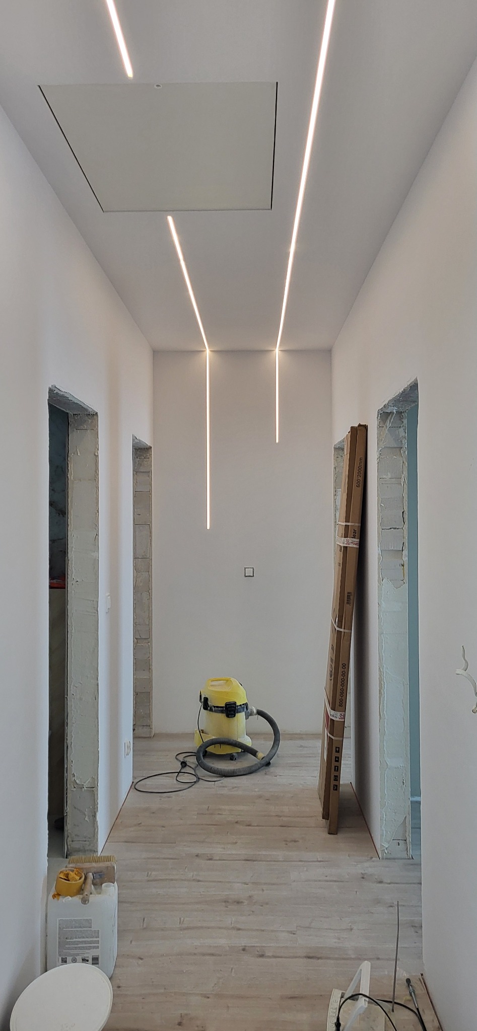 Korytarz w trakcie prac, widoczne oświetlenie ledowe w suficie, przygotowane ościeżnice drzwiowe, odkurzacz przemysłowy i materiały budowlane na podłodze.