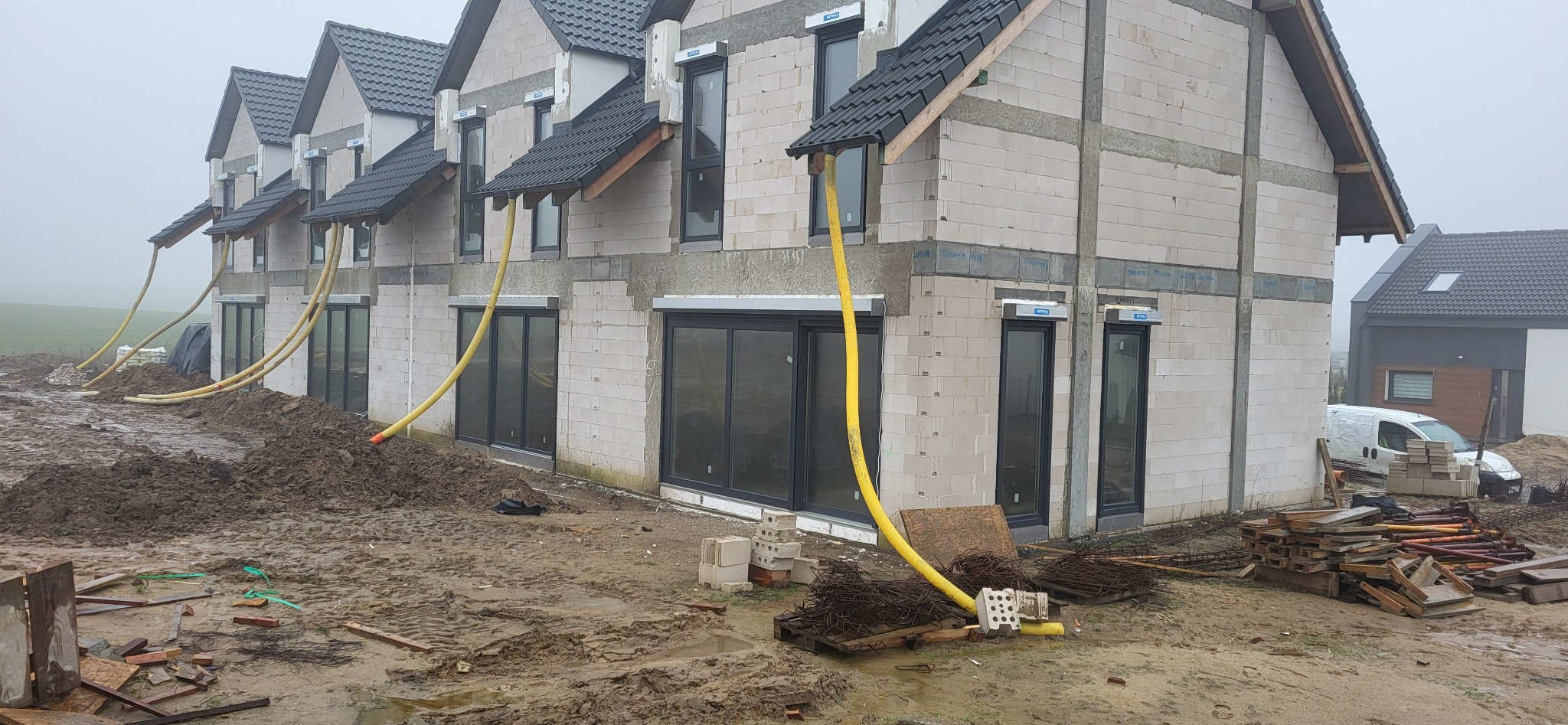 Surowy stan budowy szeregowca z białych bloczków, widoczne żółte węże wentylacyjne wystające z okien, błotniste podłoże, materiały budowlane i biały samochód dostawczy w tle.