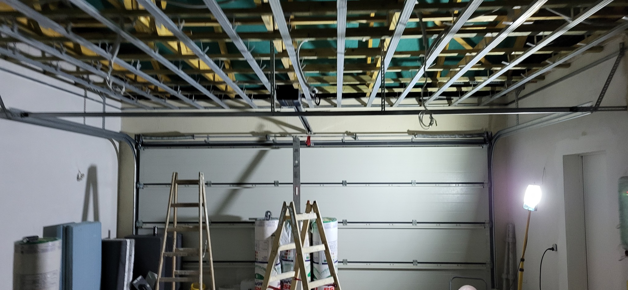 Montaż metalowej konstrukcji podwieszanego sufitu w garażu, widoczne drabiny, materiały izolacyjne i lampa robocza.