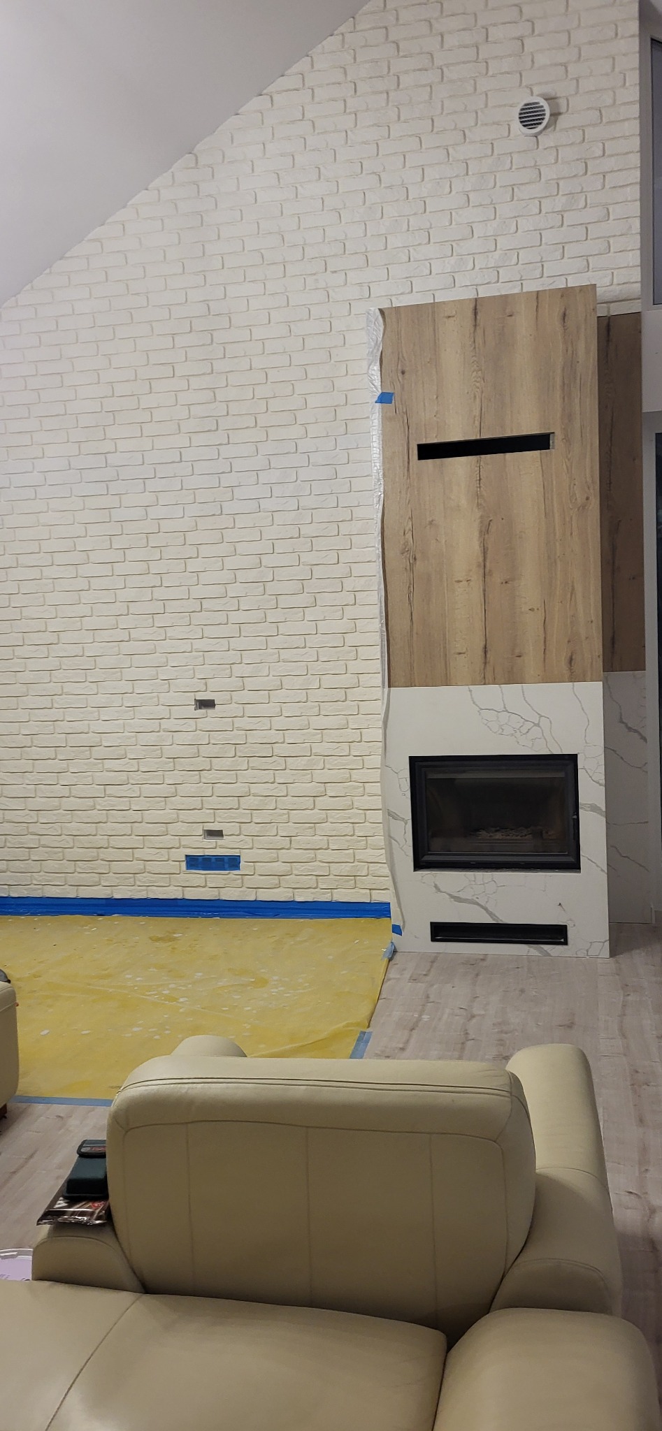 Ściana z cegły pomalowanej na biało, z zainstalowanym nowoczesnym kominkiem obudowanym drewnem i marmurem, podłoga zabezpieczona folią malarską, fragment skórzanej sofy w dolnej części kadru.