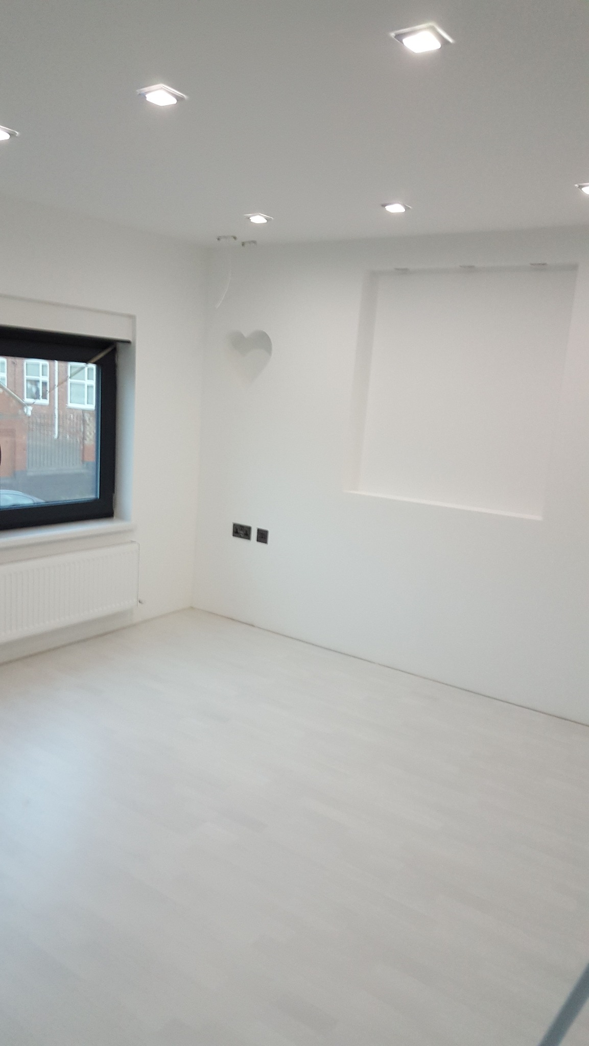 Puste, białe wnętrze po remoncie z jasną podłogą, czarną ramą okienną, białym grzejnikiem, wnęką w ścianie i dekoracyjnym sercem nad gniazdkami.