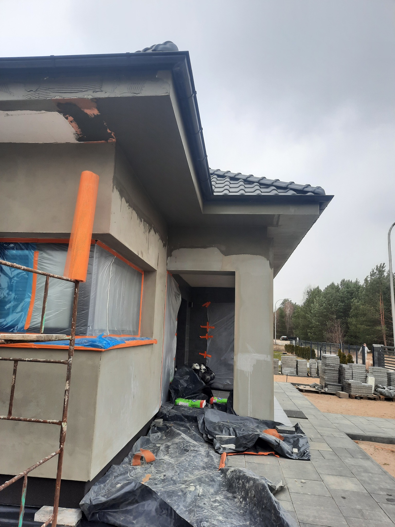 Surowa elewacja domu w trakcie prac wykończeniowych, okna zabezpieczone folią, widoczne rusztowanie i materiały budowlane na zewnątrz, świeżo położona kostka brukowa.