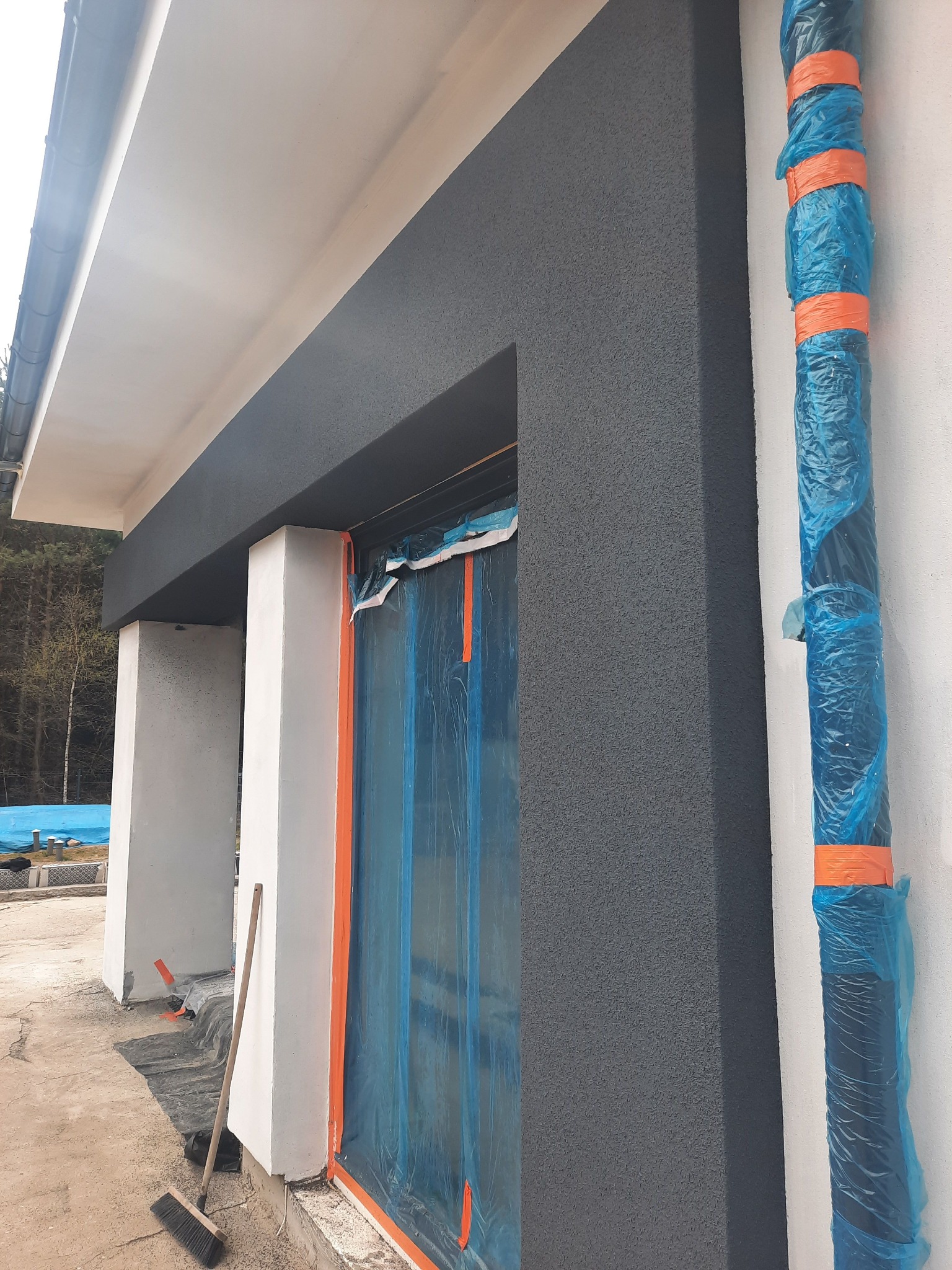 Fragment elewacji budynku z oknem zabezpieczonym folią, ściana pokryta szarą strukturą, rura spustowa owinięta folią ochronną, widoczny fragment dachu i otoczenia.