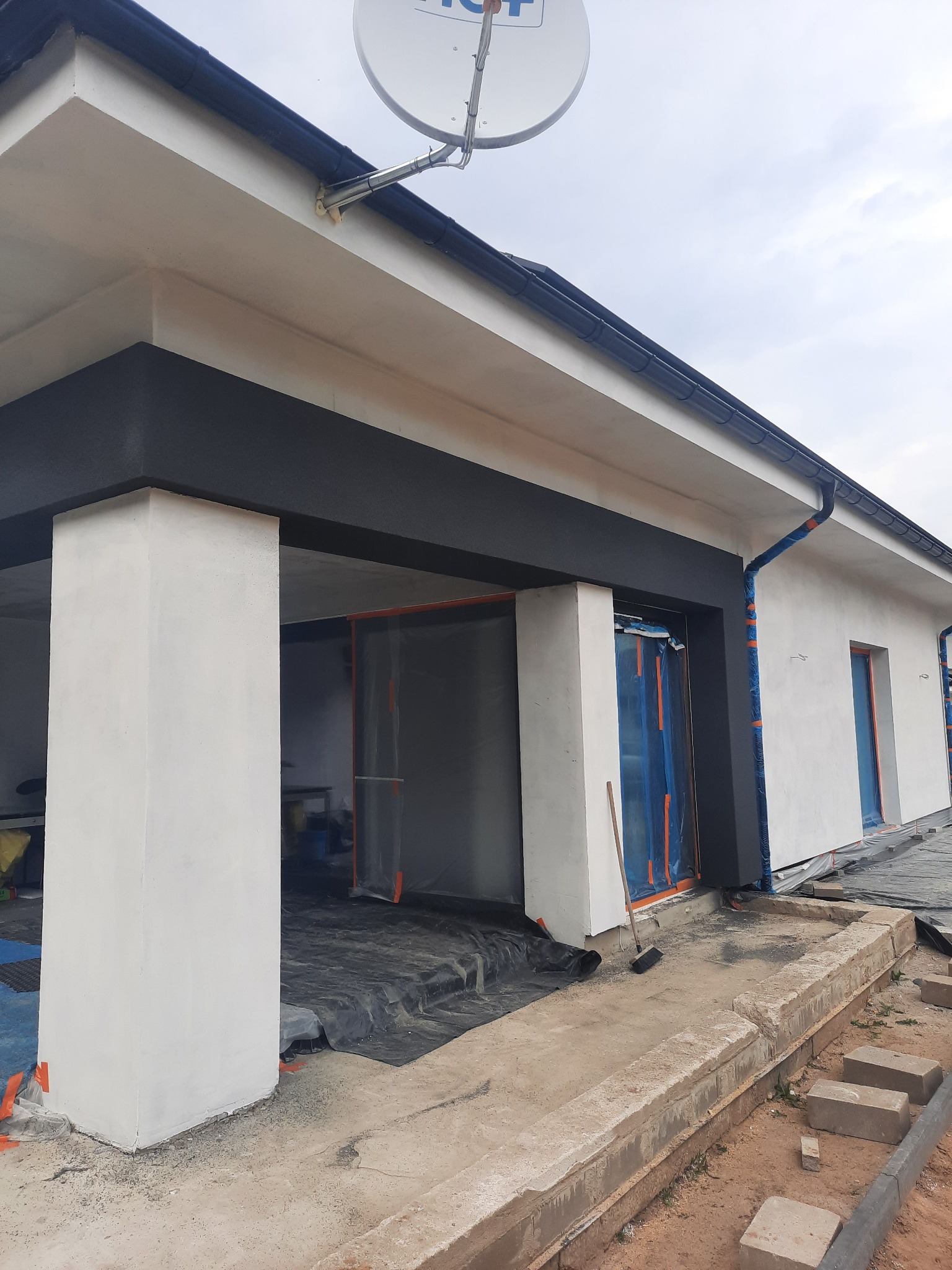 Wykończona elewacja domu jednorodzinnego z czarnymi elementami dekoracyjnymi, okna i drzwi zabezpieczone folią ochronną, widoczna antena satelitarna na dachu.