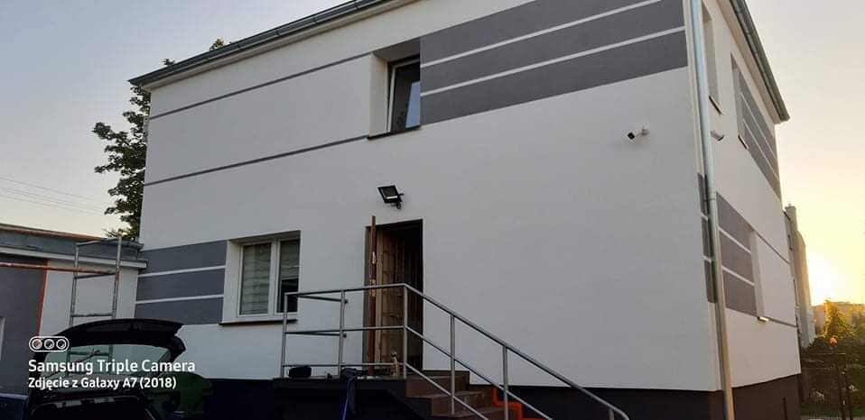 Dwukondygnacyjny budynek z białą elewacją ozdobioną szarymi pasami, widoczne schody z metalową barierką prowadzące do drzwi wejściowych.