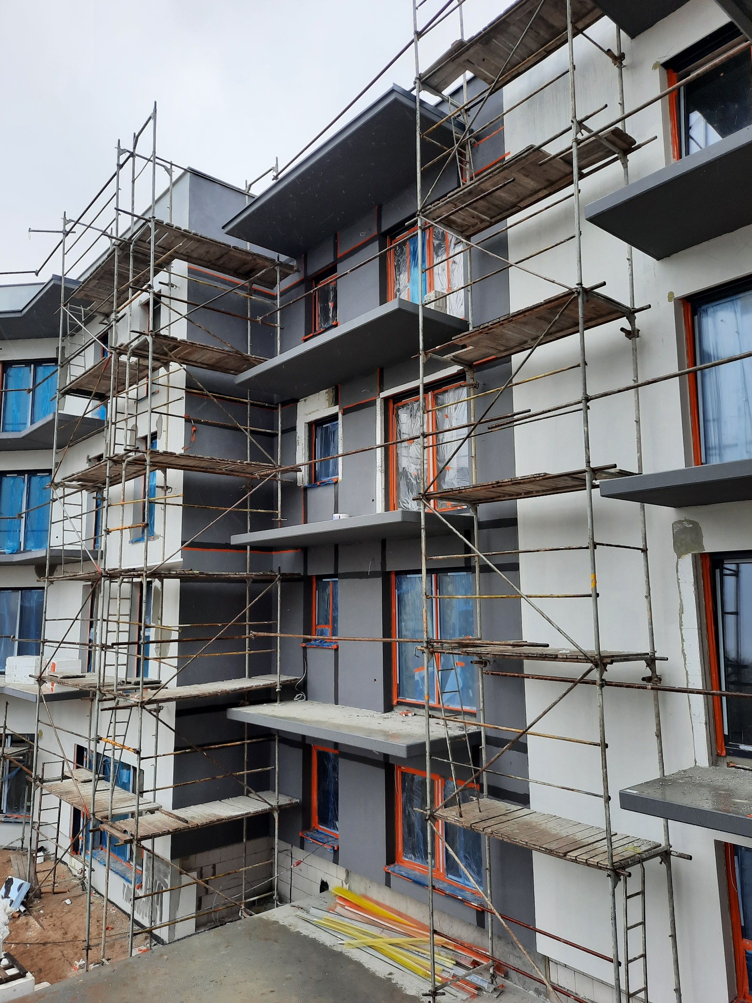 Budynek wielorodzinny w trakcie prac elewacyjnych, z widocznymi rusztowaniami, oknami zabezpieczonymi folią i fragmentami ściany w kolorze szarym i białym.