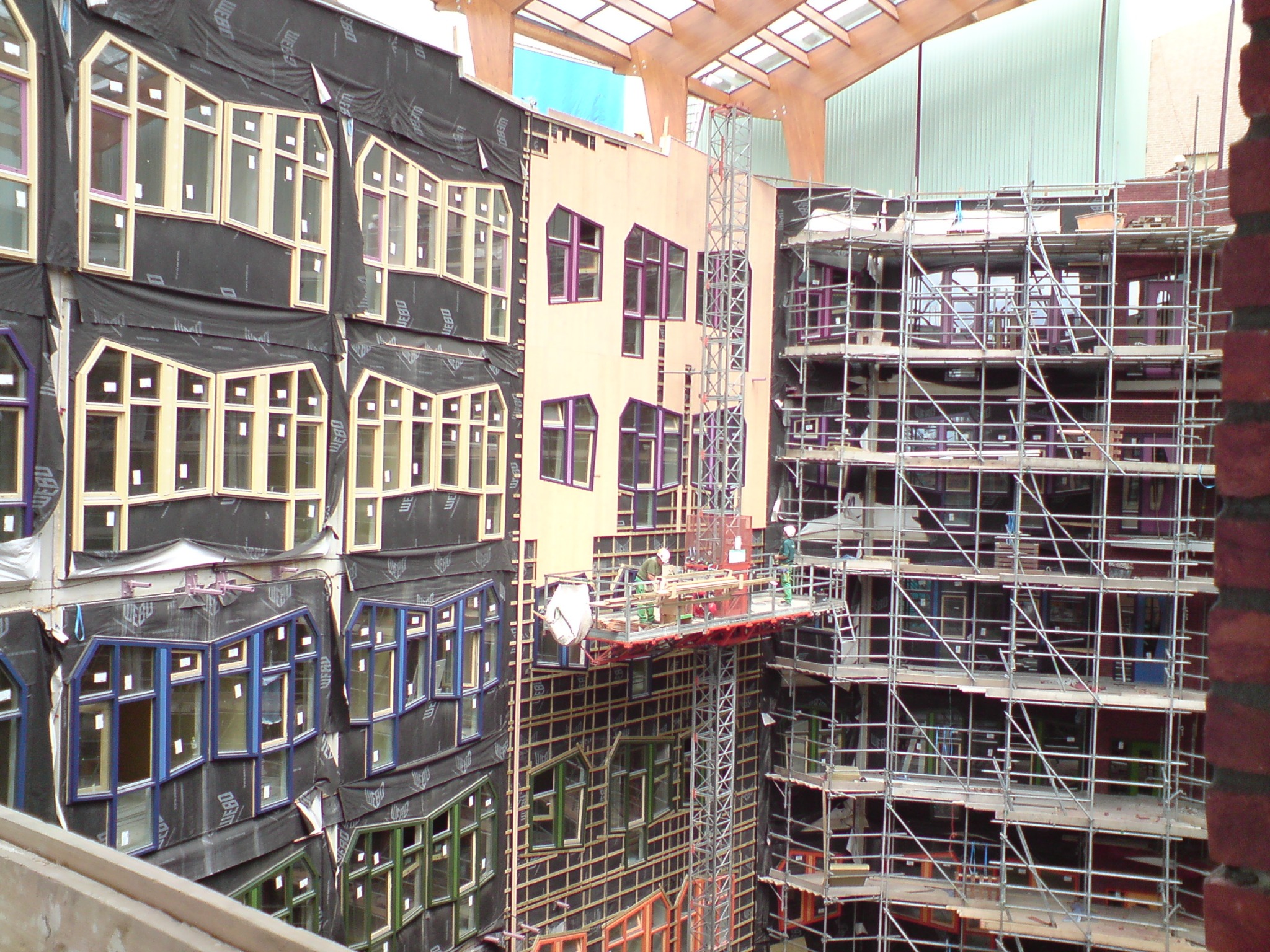 Budynek w trakcie prac elewacyjnych, widoczne rusztowania, okna o nietypowych kształtach i kolorach, ekipa robotników na podnośniku.