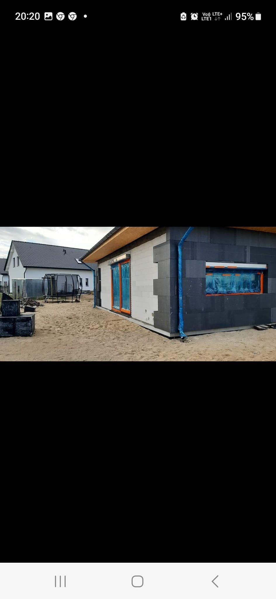 Budowa domu jednorodzinnego z szarych i białych bloczków, z zamontowanymi oknami zabezpieczonymi folią, widoczne są również rury spustowe i trampolina w tle.