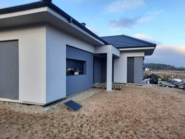 Nowoczesny dom jednorodzinny z elewacją w odcieniach szarości i bieli, widok z zewnątrz, teren wokół domu w trakcie prac wykończeniowych, widoczne palety i materiały budowlane.