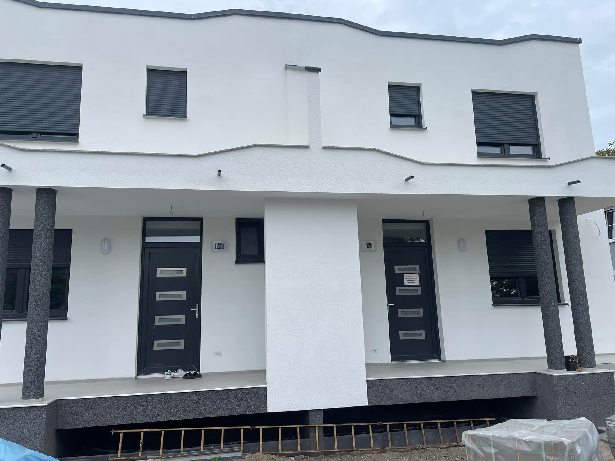Biała elewacja dwukondygnacyjnego bliźniaka z szarymi roletami i kolumnami, widoczne numery domów 123b i 125, drabina i materiały budowlane leżące przed budynkiem.