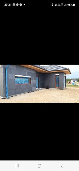 Dom jednorodzinny w trakcie ocieplania elewacji, widoczne szare płyty izolacyjne i zabezpieczone okna folią ochronną, teren wokół domu piaszczysty.