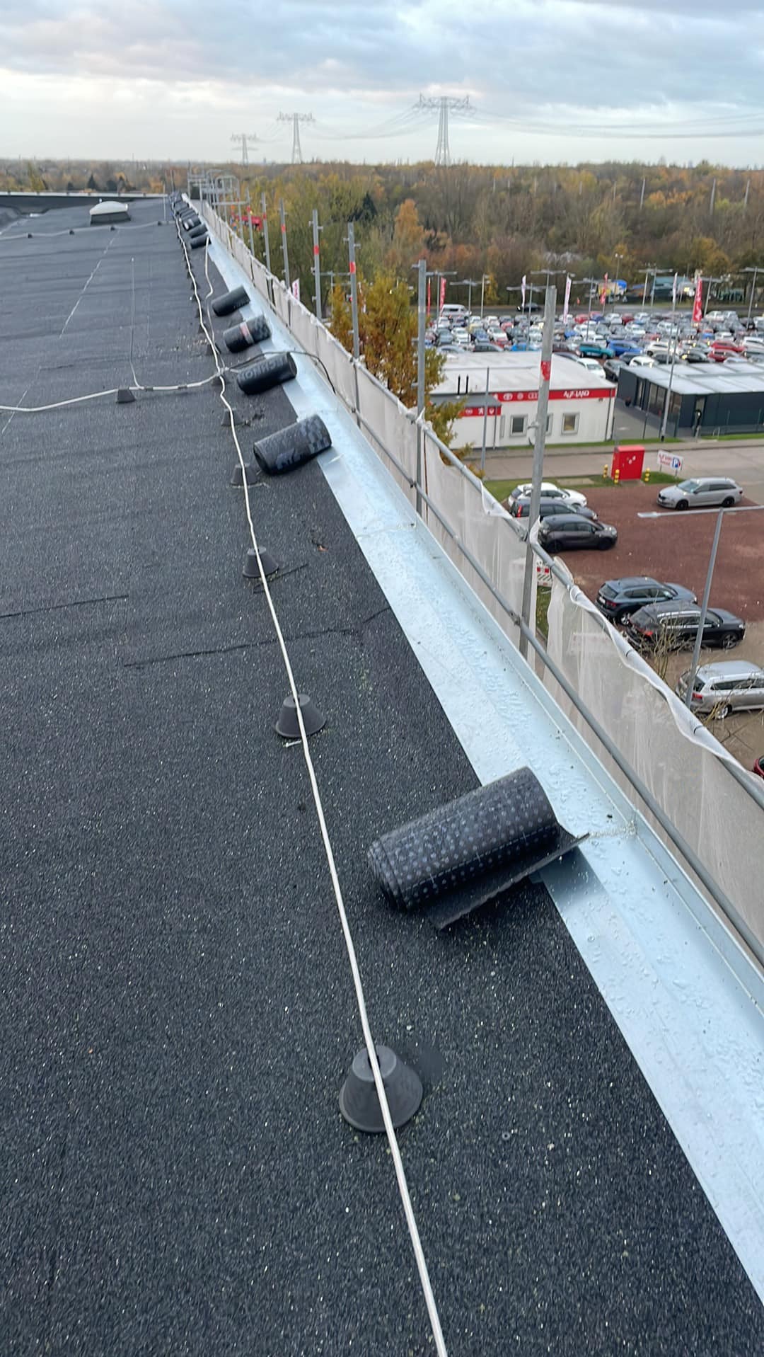 Czarny, płaski dach pokryty papą z widocznymi rolkami papy i elementami mocującymi, w tle budynki i parking.