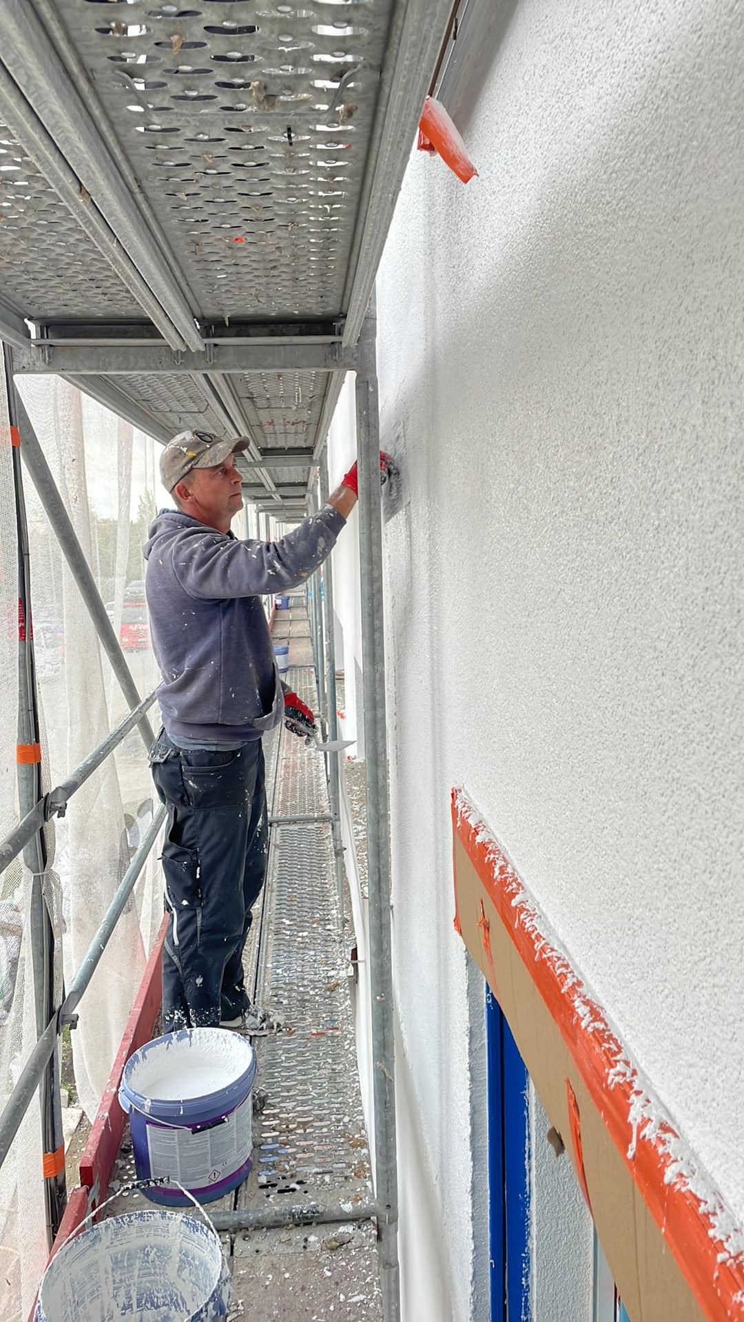 Pracownik w Pile, stojąc na rusztowaniu, nakłada białą strukturę na elewację budynku, widoczne narzędzia i wiaderka z farbą.