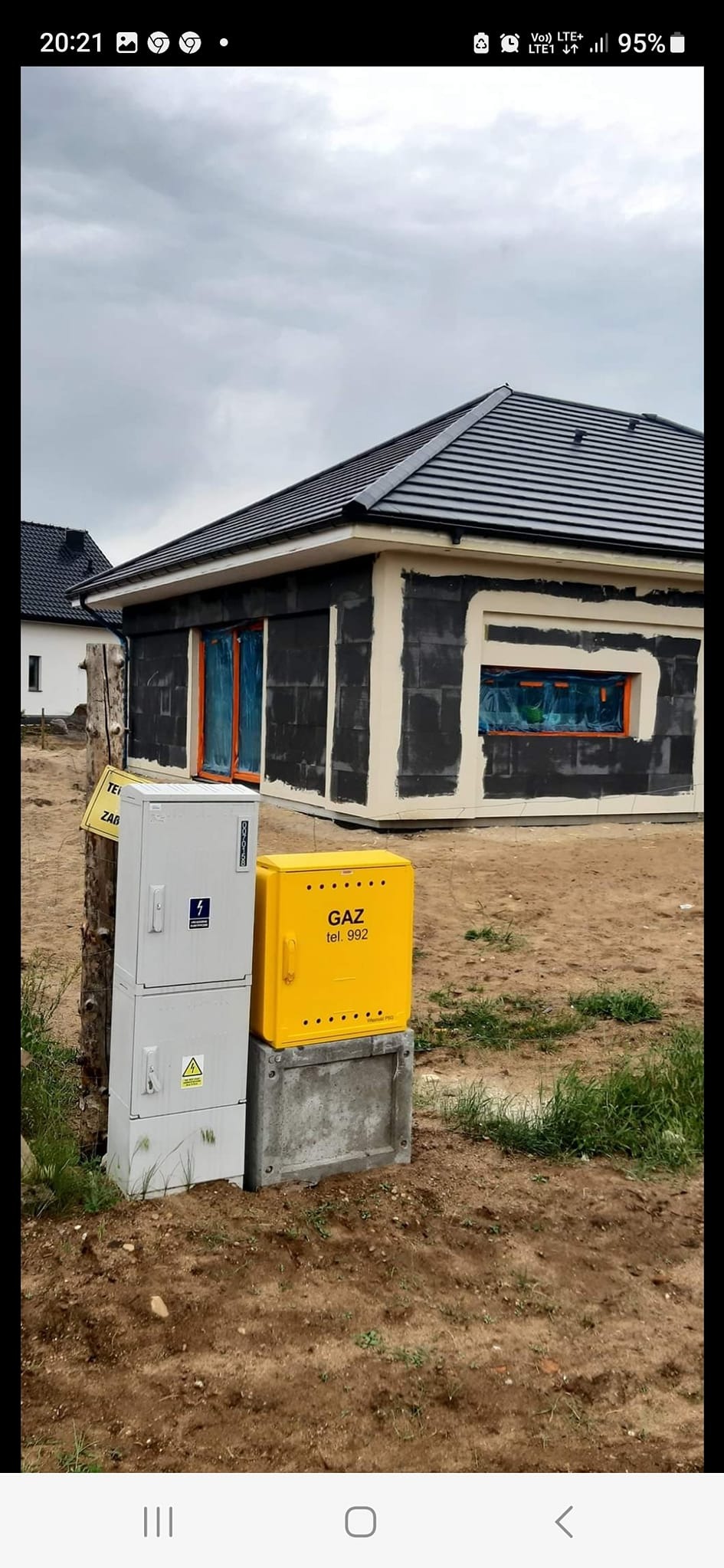 Niedokończony dom jednorodzinny z czarną dachówką, ocieplony styropianem i przygotowany do elewacji, z widocznymi skrzynkami elektryczną i gazową na pierwszym planie.